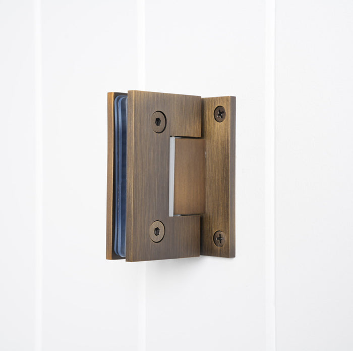 Rennes Shower Hinge Glass to Wall in Antique Brass, three-quarter view with clamped glass on a white vertical-panelled wall