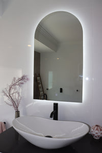 Curved Backlit LED mirror with White Vessel Sink, Black Countertop, and Faucet in White Bathroom