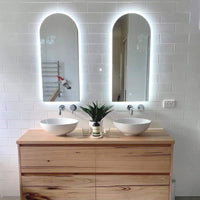 Two lighted curved backlit LED mirrors with brown cabinet, white vessel sinks on white subway tiles