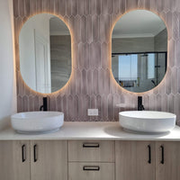 Dual Yellow-Lit Oval Smart LED Mirrors in Couple's Bathroom, Picket Tiled Wall, White-Brown Cabinet