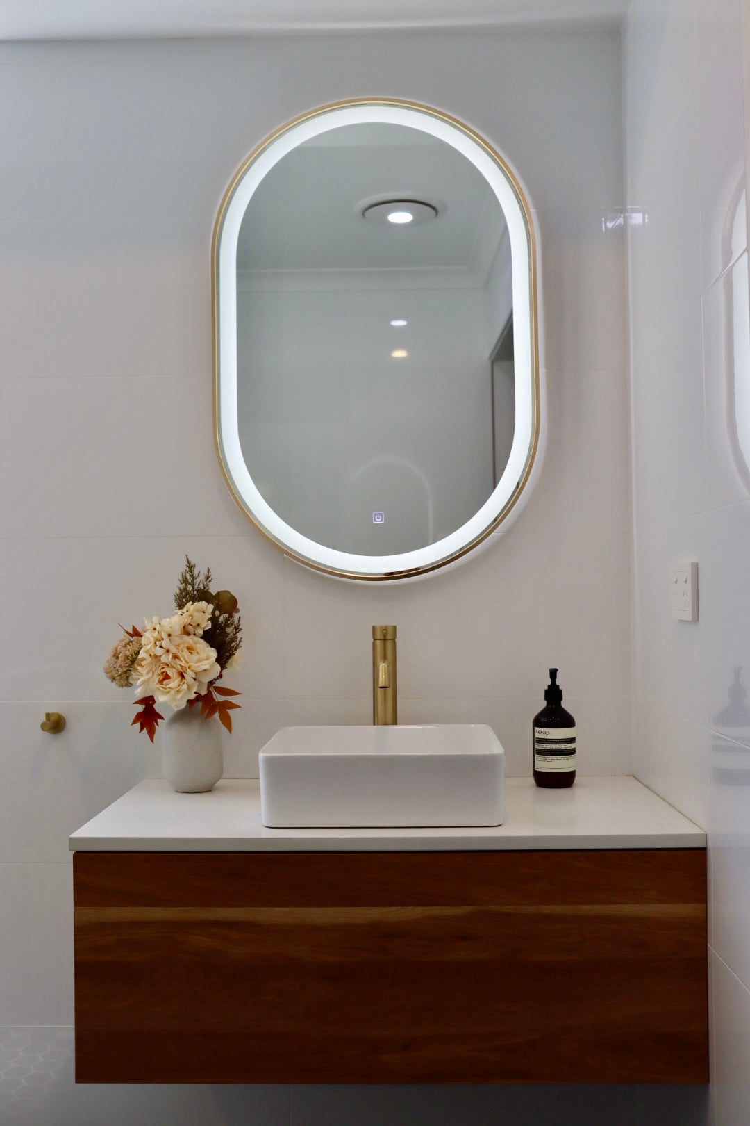 Pill-shaped LED Mirror with Silver Frame Above Floating Brown Vanity Cabinet in White Bathroom
