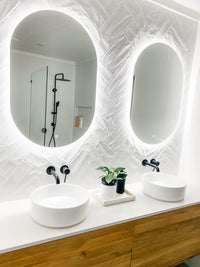 Bright vanity area with dual Oval LED mirrors and floating cabinet on white herringbone tiled wall